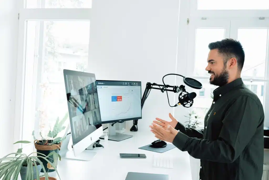 A man hosts a webinar in a white room, speaking into a microphone and gesturing for the audience online.
